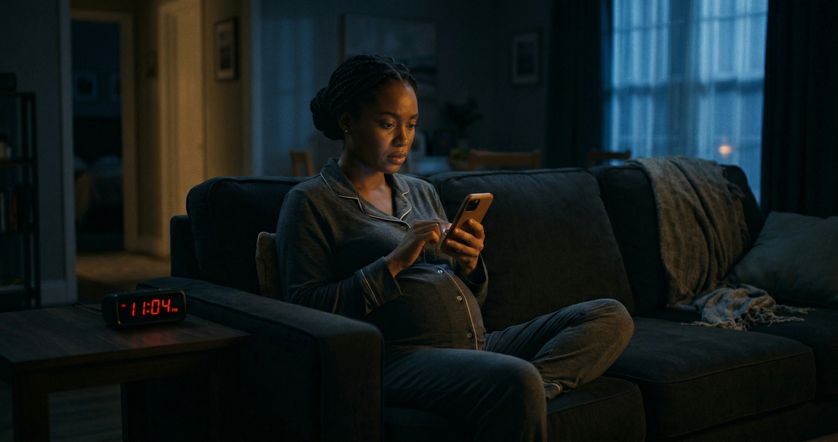 Pregnant woman sitting alone on a dark couch late at night, her face lit by the glow of her phone as she types a medical question into a chatbot